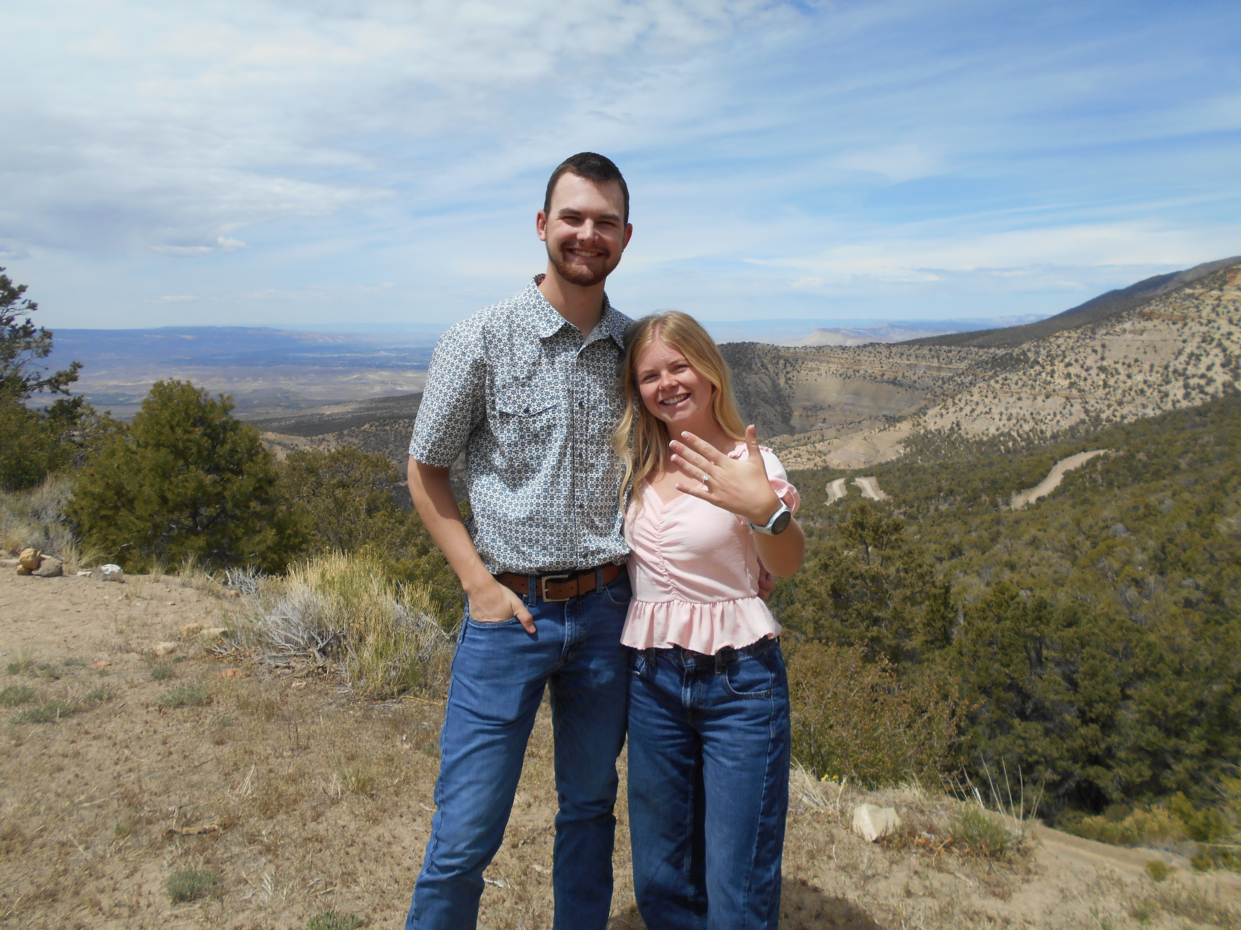 Asher and Mia smiling together at a scenic overlook, showing their engagement ring